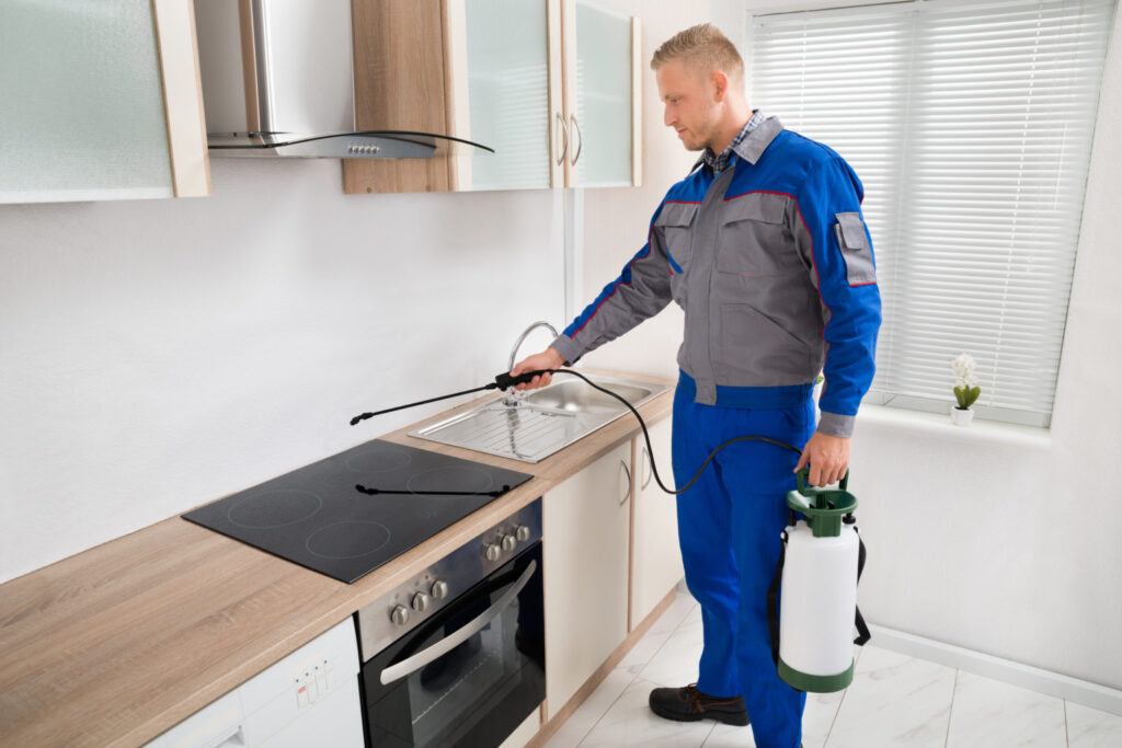 A man wearing a blue uniform and holding a white bottle, standing in a kitchen. He use his other hand to spray something from a pesticide container onto the countertop. He is performing pest control.