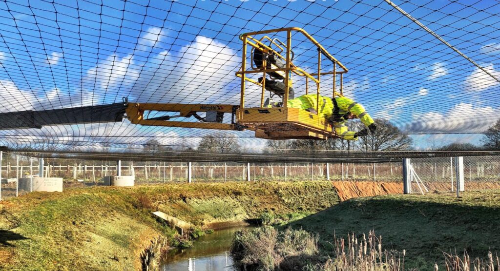 A large yellow crane at a construction site, with a blue sky in the background. There are plants, grass, and a fence around the area.