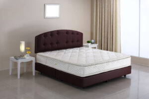 A bed in a room, showcasing interior design and furniture such as linens, pillows, bedding, and a nightstand. The room also features a window with curtains and a white color scheme.