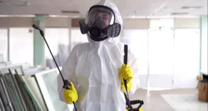a woman wearing a mask and yellow gloves. She is indoors and standing next to a wall. The tags include clothing, personal protective equipment, helmet, hazmat suit, gas mask, workwear, and wearing.